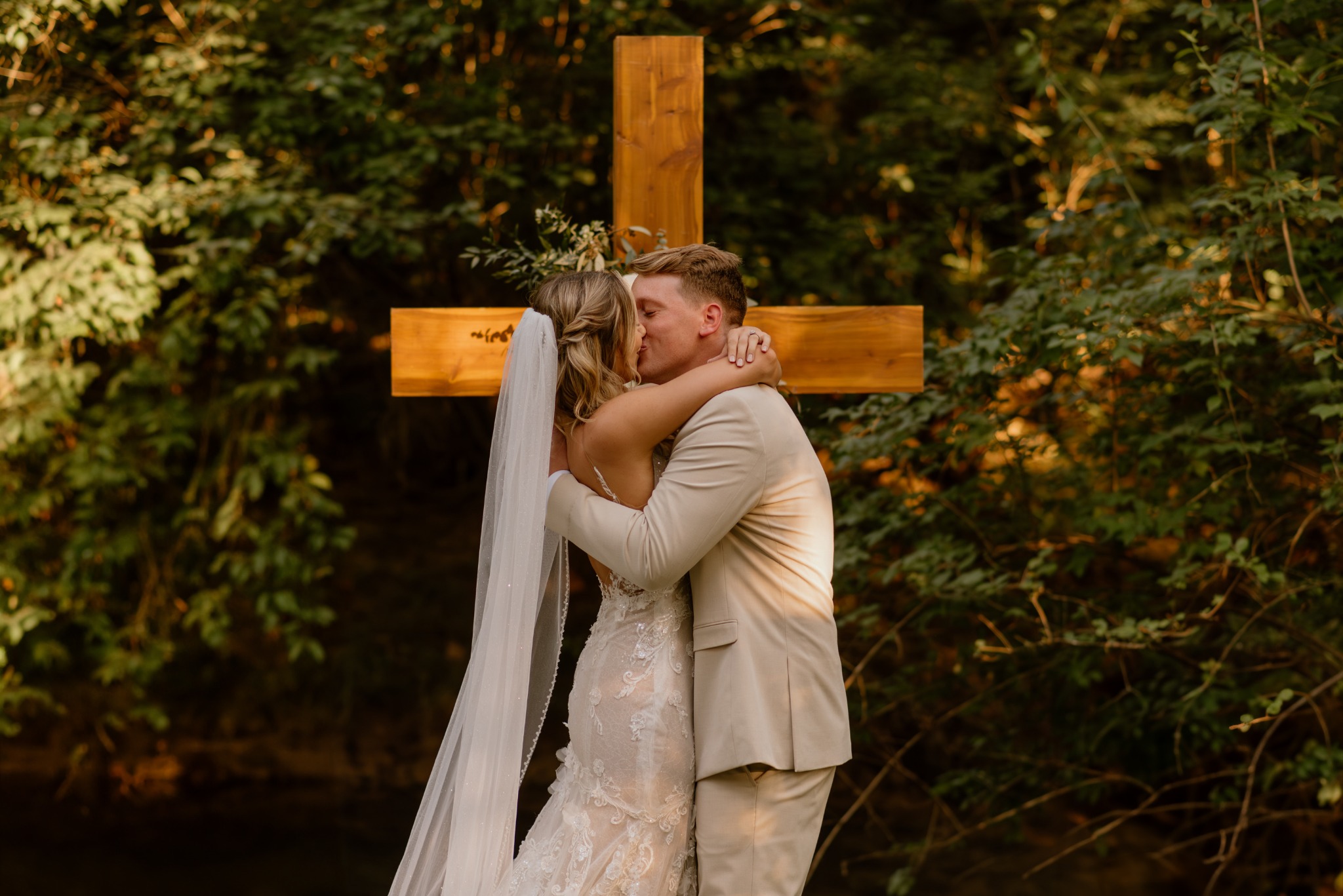 Newlyweds sharing a kiss in front of the wooden cross surrounded by the estate woods