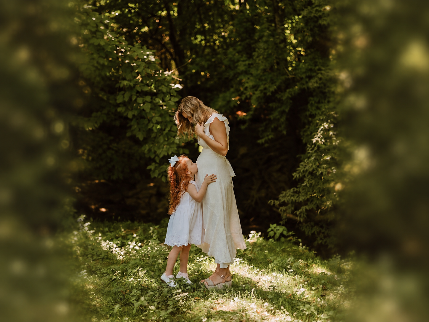 An expectant mother and her young daughter sharing a quiet portrait in a sunlit garden clearing